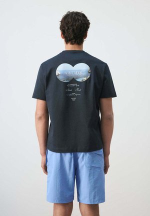 Hombre de espaldas que lleva una camiseta negra con estampado de vista al mar en forma de binoculares y texto, combinado con pantalones cortos azul claro sobre un fondo liso.