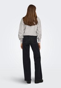 Femme aux longs cheveux bruns portant un sweat-shirt gris clair, un jean noir à jambes larges et des chaussures noires, vue de dos sur un fond uni.