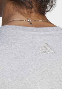 Světle šedé bavlněné tričko s žebrovaným výstřihem a vyšitým logem Adidas na zadní straně. Obsahuje jemný stříbrný náhrdelník.