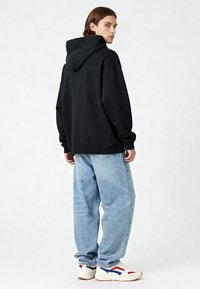 Čierna mikina s kapucňou vo voľnom strihu, skombinovaná s svetlomodrými baggy džínsami a bielymi teniskami s červenými a modrými akcentmi.
