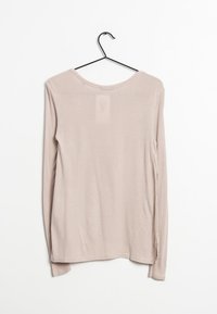 Camicia beige a maniche lunghe appesa a un gancio nero, con scollo rotondo e una texture morbida e liscia, con un pannello posteriore leggermente trasparente.