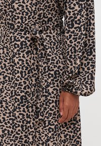 Haljina s leopard uzorkom koja sadrži crne uzorke na bež pozadini, pojas u struku, nabrane rukave i teksturiranu tkaninu.