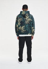 Sudadera con capucha de patrón de hojas verde y beige, sobre un fondo negro. Combinada con pantalones cargo negros y zapatillas blancas.