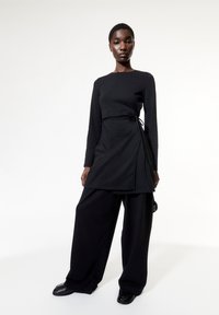  långärmad svart topp med en omslagsdesign och knytning i midjan, tillsammans med vida svarta byxor och släta svarta skor.