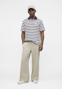 Άνδρας όρθιος με τα χέρια στις τσέπες, φορώντας λευκή και ναυτική ριγέ πόλο μπλούζα, μπεζ παντελόνια με φαρδιά πόδια, κρεμ παπούτσια και μπεζ καπέλο.