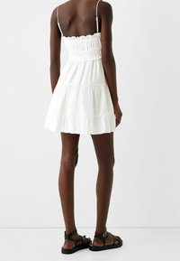 Kvinna klädd i en ärmlös vit tiered minikjol med smockad baksida och tunna axelband, matchat med svarta platta sandaler med ankelremmar.