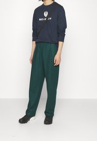 Man som bär en marinblå långärmad skjorta med vit logotyp, mörkgröna lösa byxor, svarta skor och en svart axelrem.