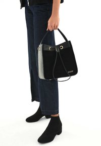 Bolso tipo cubo en negro y gris con herrajes dorados, con textura de ante y cierre de cordón, sostenido por una persona que lleva jeans oscuros y botas negras.