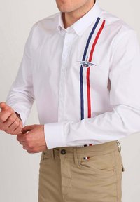 Homme portant une chemise blanche à boutons avec des rayures verticales rouges et bleues et un emblème d'avion, associée à un pantalon beige.