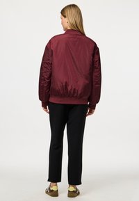 Burgundy bomber jacket with ribbed collar and cuffs, shiny texture; paired with black pants and brown sneakers with yellow accents.
