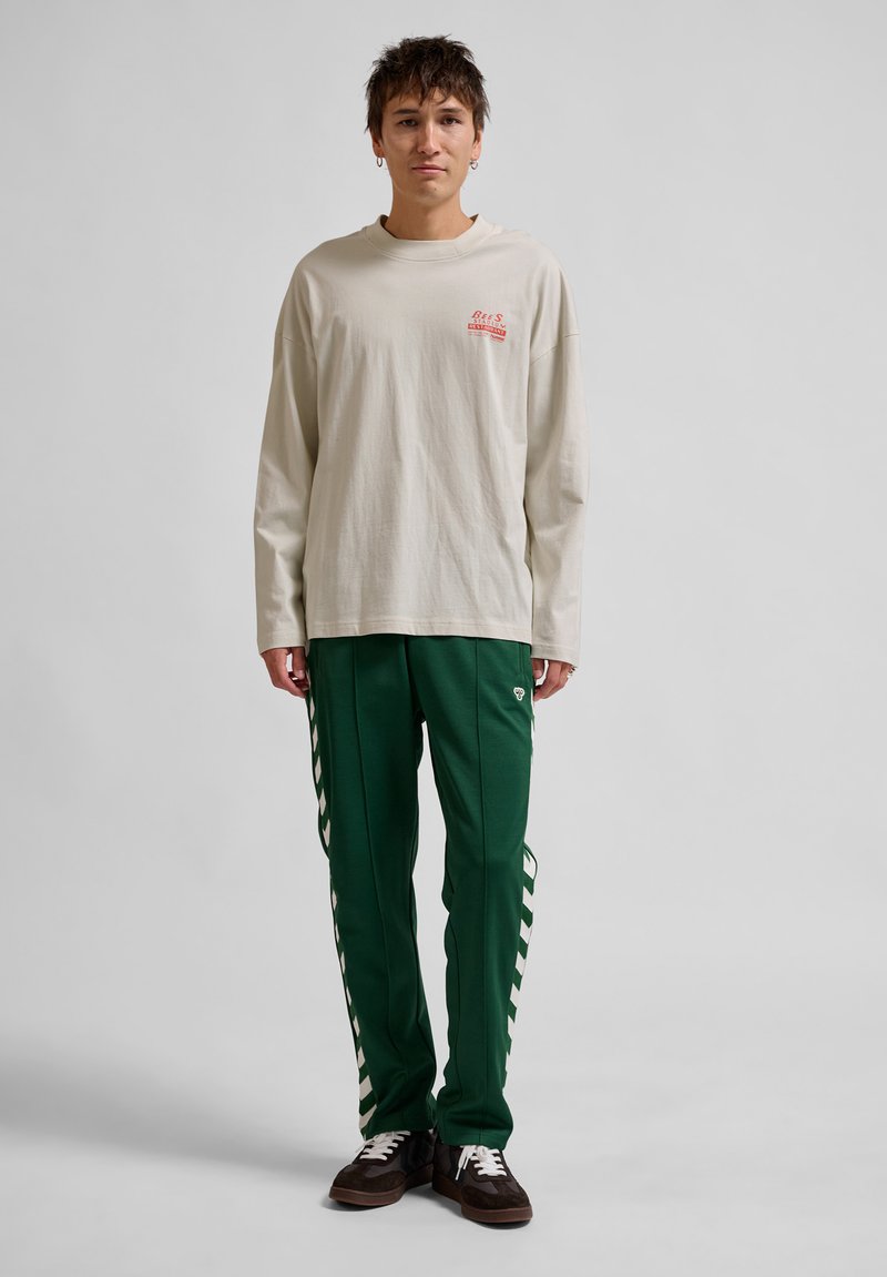 Cremefarbendes Langarmshirt mit roten grafischen Akzenten, kombiniert mit grünen Sporthosen mit weißen Seitenstreifen. Braune Sneakers runden das Outfit ab.