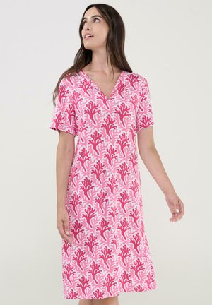 Mujer con vestido rosa de manga corta hasta la rodilla con un patrón rojo similar a coral, mirando hacia arriba contra un fondo liso claro.