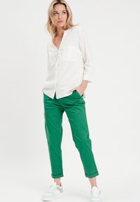 Chemise blanche à manches longues avec deux poches sur la poitrine ; associée à un pantalon vert retroussé et des baskets blanches. Fond uni.