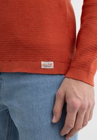Orange strukturiertes Strickpullover mit einem taillierten Saum, versehen mit einem kleinen Etikett am unteren Ende. Getragen mit hellblauen Jeans.