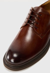 Scarpa stringata in pelle marrone con finitura liscia, punta rotonda e suola scura a contrasto. Presenta sei fori per i lacci e dettagli cuciti.