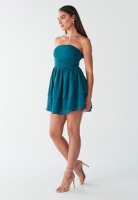 Teal, strapless mini-jurk met een aansluitend lijfje en een gelaagde, uitlopende rok. Draagt gouden, strapless hoge hakken.