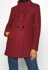 Rød frakke i uldblanding med tekstureret finish, dobbeltradet design, sorte knapper, sidelommer og høj krave.