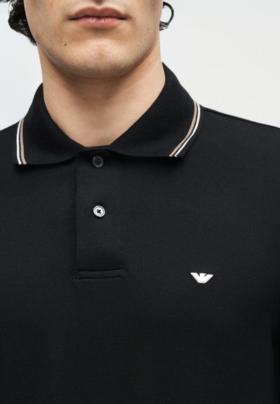 Άνδρας που φοράει μαύρο πόλο πουκάμισο με ριγέ γιακά και μικρό λευκό λογότυπο στο στήθος, ορατός από το πηγούνι έως την άνω πλευρά του κορμού.