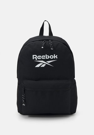 Le sac à dos noir présente un design robuste, un logo Reebok blanc bien visible, des bretelles réglables et une poche zippée à l'avant avec des tirettes décoratives.