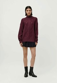 Woman wearing a burgundy cable-knit turtleneck sweater, black pleated skirt, and black ankle boots, standing against a plain background.