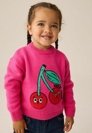 Junges Kind mit geflochtenem Haar, das einen leuchtend pinken Pullover trägt, auf dem zwei lächelnde Kirschen abgebildet sind, sowie eine blaue Jeans.