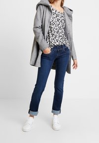Manteau à capuche gris porté sur un haut à motifs noir et blanc, associé à un jean bleu foncé et des baskets blanches. Texture douce et coupe décontractée.