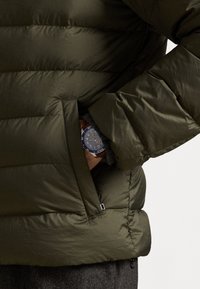 Geacă puffer de culoare verde închis, cu un design matlasat, buzunar lateral și mansetă de pulover gri vizibilă. Un ceas albastru este parțial vizibil.