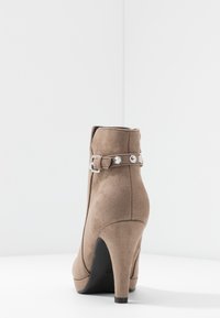 Brown suede ankle boot with a high heel and a decorative belt featuring silver hardware details. Smooth texture with a rounded toe design.