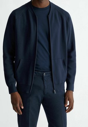 Man wearing navy blue zip-up jacket over matching navy t-shirt and pants, standing with hands relaxed at sides.