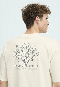 Lys beige bomulds t-shirt med et grafisk tryk af en bomuldsplante med stiliserede blomster på ryggen. Korte ærmer og en afslappet pasform.