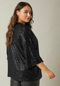 Top en sequins noirs avec un design ample et des manches chauve-souris, présentant une surface texturée et des manches larges. Les sequins brillants reflètent la lumière.