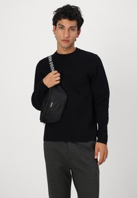 Schwarze Umhängetasche mit glatter Textur, ausgestattet mit einem kontrastierenden weißen Tragegurt mit der Aufschrift "HUGO". Einfache Reißverschlussöffnung und elegantes Design.