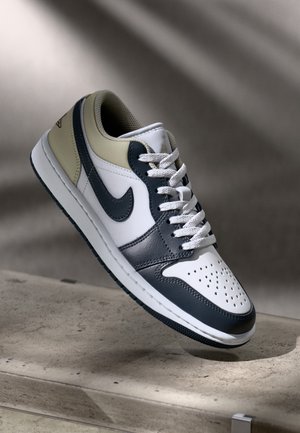 Białe, czarne i beżowe niskie sneakersy Nike z perforowanym przodem, czarnym logo Swoosh, białymi sznurówkami, przedstawione unoszące się nad kamienną powierzchnią.