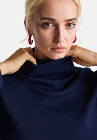 Maglione a collo alto blu navy realizzato in morbido tessuto a maglia, con colletto ripiegato e cuciture visibili. Il modello si aggancia al collo con le mani.