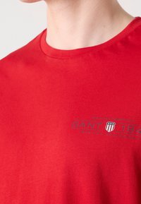 T-shirt rouge à col rond avec petit texte « GANT 1949 » et logo bouclier bleu sur la partie supérieure de la poitrine.