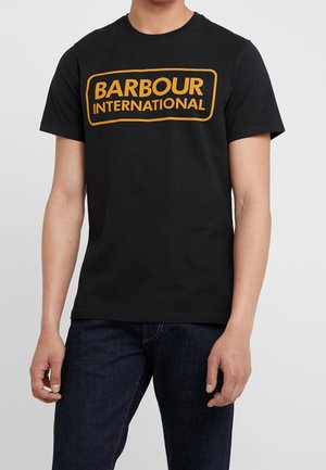 Mann trägt ein schwarzes T-Shirt mit kurzen Ärmeln und gelbem „BARBOUR INTERNATIONAL“-Schriftzug, kombiniert mit dunkelblauen Jeans, steht vor einem einfarbigen Hintergrund.