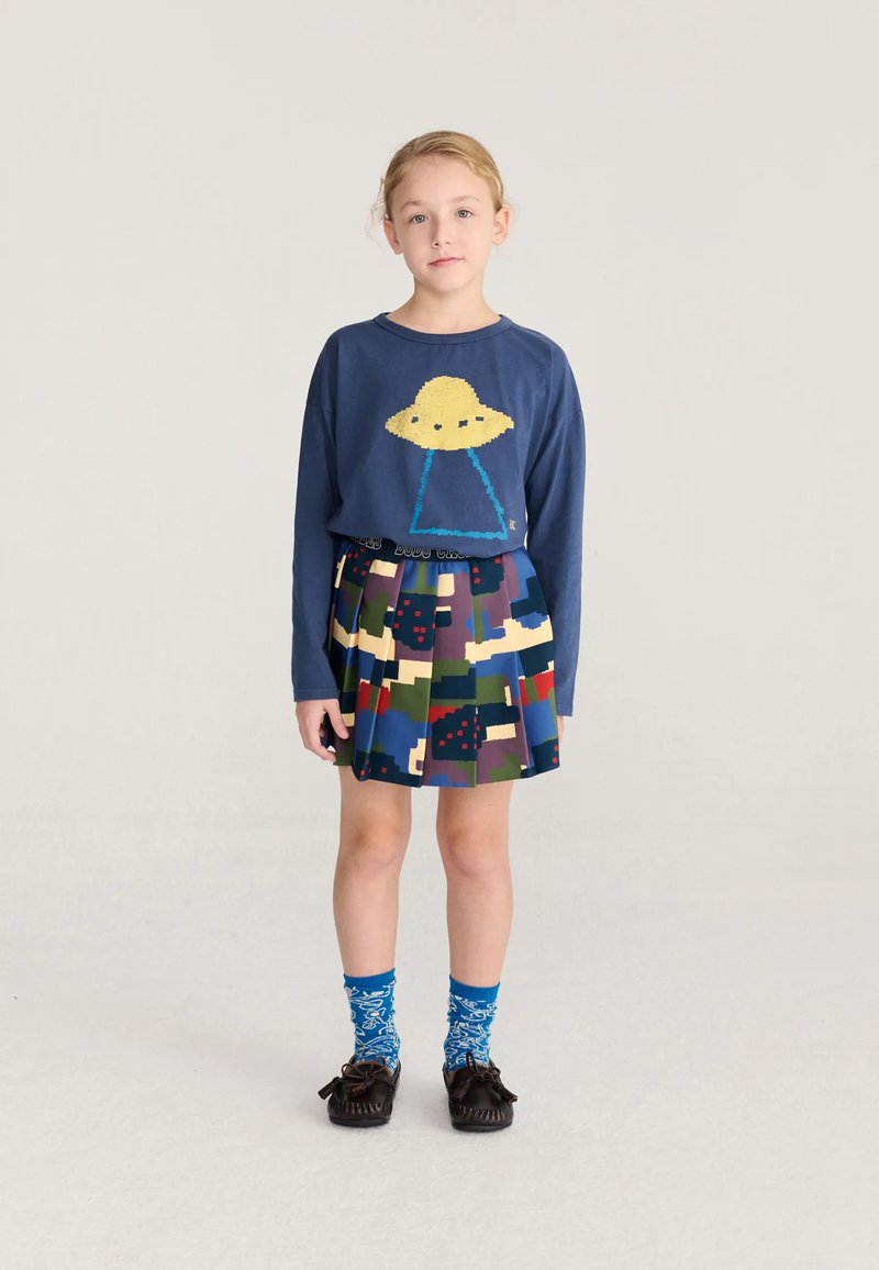 Enfant aux cheveux blonds debout, portant un sweat-shirt bleu marine avec un OVNI jaune, une jupe multicolore, des chaussettes bleues à motifs et des mocassins foncés.