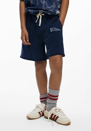 Bambino che indossa pantaloncini blu navy con tasca frontale e patch "Desigual", calzini grigi con strisce rosse e sneakers bianche con dettagli rossi.