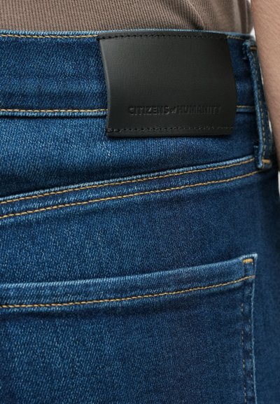 Kék farmernadrág fekete bőr derékpánttal, amelyen a „CITIZENS of HUMANITY” felirat látható. Narancssárga varrással és hátul zsebfedővel rendelkezik.