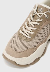 Sneaker beige realizzata in materiali mesh e suede. Presenta lacci crema, dettagli testurizzati e un logo rotondo sulla linguetta. Suola in gomma spessa.