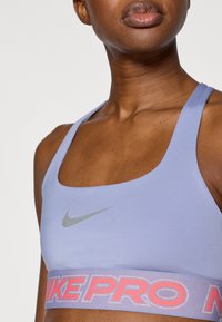 Αθλητικό σουτιέν Nike Pro σε απαλό μωβ χρώμα, με γκρι λογότυπο και ευρύ, υφασμένο λάστιχο στο κάτω μέρος που φέρει τη φράση "NIKE PRO" σε ροζ και λευκό.