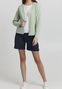 Ljusgrön cardigan med ett mönster, bärs öppen över en vit skjorta, matchad med marinblå shorts och vita sneakers.
