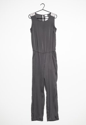 Combinaison grise en tissu lisse, dotée d'une taille cintrée, d'un design sans manches et d'accents décoratifs aux épaules. Accrochée à un crochet noir.