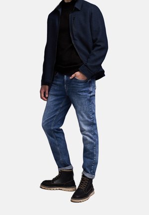 Blaue Jeans aus Denim, schwarzer Pullover und dunkelblaue Jacke; schwarze Stiefeletten mit beige farbenen Sohlen; Reißverschluss-Detail an der Jacke; lässige Passform.