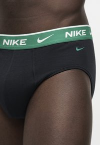 Gros plan d'une personne portant un slip Nike noir avec une ceinture verte affichant des inscriptions et logos Nike blancs.