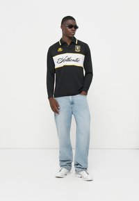 Polo a maniche lunghe nera con accenti bianchi e gialli, con la scritta "Authentic". Un paio di jeans azzurri e sneakers bianche.