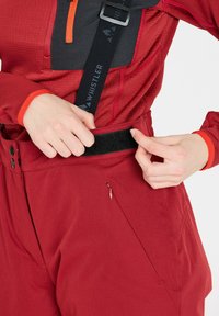 Röda utomhusbyxor med en slät yta, justerbara remmar och en sidoficka med dragkedja; utrustade med ett kardborreband och kontrasterande mörka detaljer.