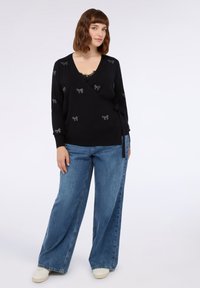 Svart omslagströja med liten silverfärgad rosettbroderi, tillsammans med lösa blå denimjeans och vita sneakers. Slen struktur, långa ärmar, knytdetalj.