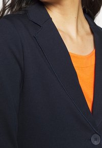 Donna che indossa un blazer blu scuro sopra un top arancione brillante, mostrando la clavicola e i dettagli dei bottoni del blazer.