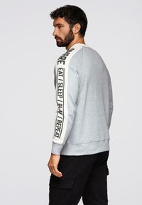Sudadera gris claro con cuello redondo, mangas largas que presentan una banda de crema contrastante con texto repetido y dobladillo acanalado.